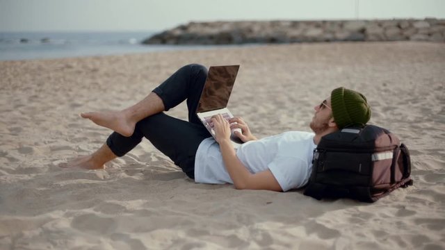 Young man in hipster millennial beanie lays on beach, relaxed barefoot in casual clothing. Works remotely from vacation spot during weekend, send and reply to emails from laptop