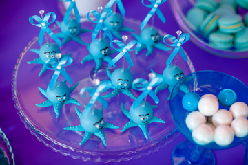 Blue cake pops funny octopuses shared on the glass round plate and jars whith marshmallow on purple background. Summer candy bar on the party, birthday