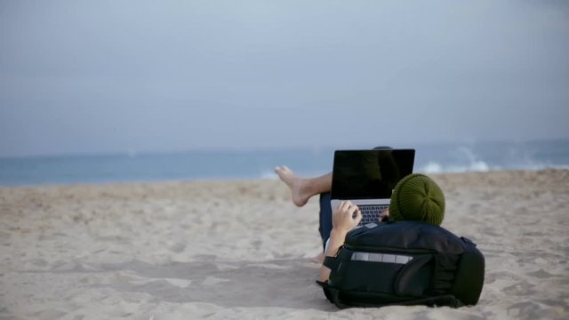 Camera moves around urban digital nomad, young entrepreneur or freelancer, millennial or generation z age, lay down at beach with backpack, work remotely on laptop, check on updates