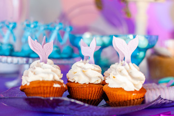 Birthday party concept for girl. Table for kids with cupcakes with withe topind decored purple mermaid tail. Summer season delicious on the party. Sea time theme on the candy bar