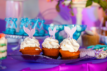 Birthday party concept for girl. Table for kids with cupcakes with withe topind decored purple mermaid tail. Summer season delicious on the party. Sea time theme on the candy bar