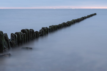 Long exposure photo of a wooden wavebreaker in the evening. Port or ship lights visible in the background.