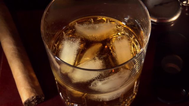 Slow motion of whiskey being stirred with ice in glass.
Cigar, cigar cutter and old pocket watch next to glass in shadows.