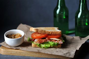 Sandwich with bacon, cheese, slices of tomatoes and green salad leaves. In the frame is a cup with mustard and two bottles of drink. The concept of a quick and healthy snack. Dark background. Close-up