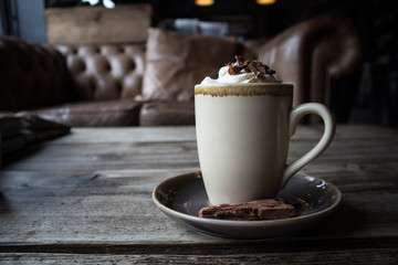 Cup of Hot Chocolate with Squrty Cream and Chocolate Sprinkles on Top.  Flake on saucer.  Standing on rustic wooden table in cafe