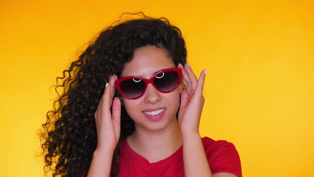 Portrait of young spanish girl with curls puts on sunglasses on yellow background. Tempting woman smiling to camera. Studio footage. Female in red t-shirt, casual style