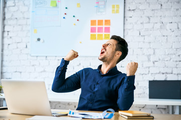 Ecstatic web developer screaming in joy and making gestures celebrating success while sitting in office - obrazy, fototapety, plakaty