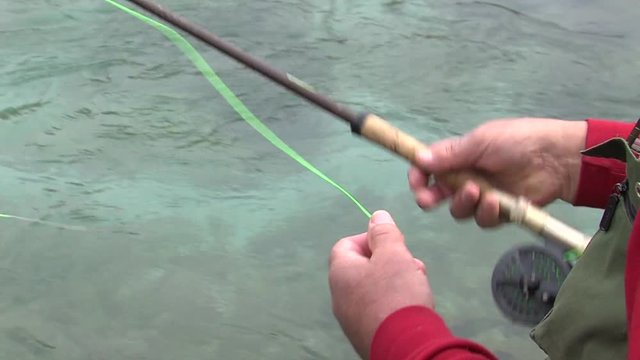 Fisherman pulling green thread then throwing fishing rod out to clean emerald river on cloudy spring day.