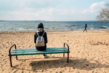 young woman traveler sits near the beach in the fall in windy weather. full length photo of girl in coat, hat, backpack and large scarf outdoors