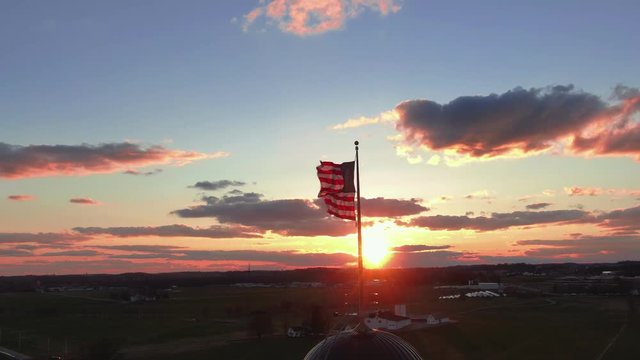 Sunset over American countryside with the American flag waving on pole and moving across the sky in front of the setting sun, dramatic skies, romatic and patriotic scene, 4k UHD
