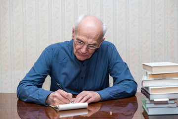 an elderly man in a blue shirt writes in a notebook with a ballpoint pen