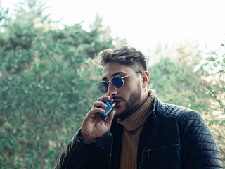 Bearded man smokes vape close up. Electronic cigarette concept. Man with long beard and clouds of smoke looks relaxed. Man with beard and mustache on calm face, branches on background, defocused