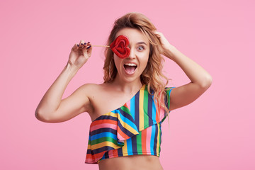 Sweet and candy. The girl colorful dress on pink background in the studio. Woman holding a red heart-shaped lollipop. Valentine's Day. Girl holding a lollipop in open mouth. Wow and shocked concept. 