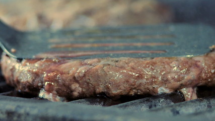 A close-up side view of a cutlet pressed with an iron spatula to the grill is fried and gives off meat juice and burrs