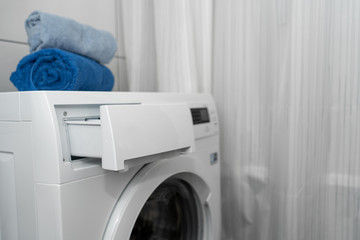 washing machine with an open box for detergents and clean folded cotton blue towels on it, in the white bathroom. Side view