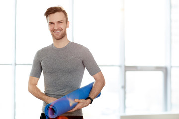 Fototapeta na wymiar Content confident handsome male yoga instructor with exercise mat standing against window and smiling at camera