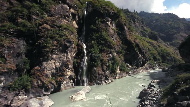 Wide angle view across fast cascading waterfall flowing into rapids creek waters below. Single cable bridge in Nepal valley trails.