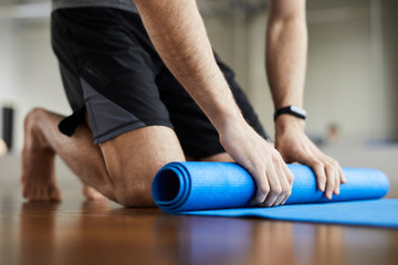 Close-up of unrecognizable man standing on knees and rolling out exercise mat after yoga training...