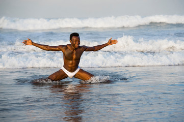 Fototapeta premium artistic portrait of young attractive and fit black African American man with sexy muscular body and six pack abs opening arms free on beach with big sea waves