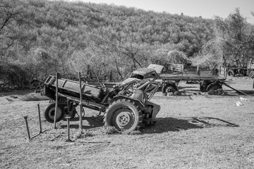 Fototapeta na wymiar Abandoned old rusty tractor, car body and farm trailers sitting in green grass field in backyard in early spring sunny day, background with leadless forest. Bulgaria, Pazardzhik region. Monochromatic