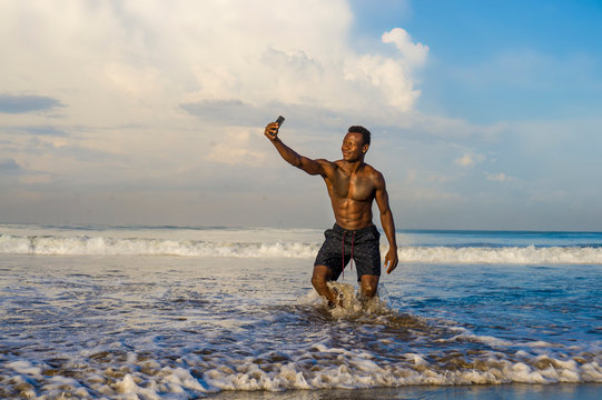 young attractive and happy black African American sport man with athletic body and sixpack taking selfie photo with mobile phone at beautiful beach smiling cool in holidays