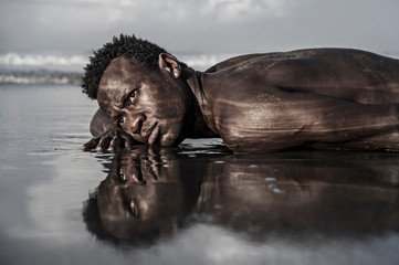 artistic expressive portrait of young attractive and sexy black African American man with athletic muscular body posing cool in sea water on desert beach in male beauty