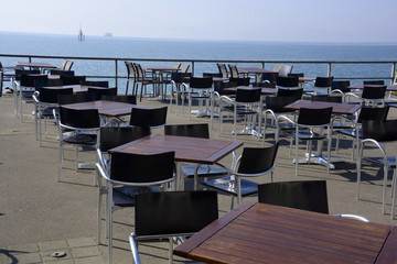 tables and chairs without persons on the lakeside promenade against lake constance and ferry and bouys at the horizon, empty restaurant in early spring at lake constance