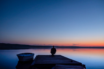 Naklejka na ściany i meble A man sits on the pier during sunset near the boat