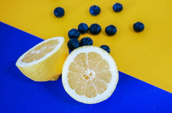 A halved lemon and a bunch of blueberries on an abstract, geometric color blocking yellow and blue background.