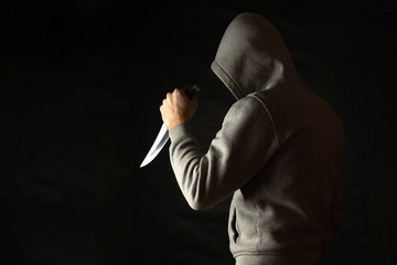 A man in a hood on a black background, brandishing with a knife in his hand. A criminal with a knife attacks from the dark.