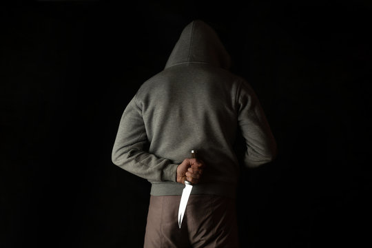 A man holds a knife in his hand behind his back, in a hood on a black background. Criminal with a knife hiding in the dark.
