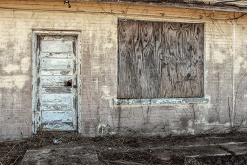 Boarded up window and vintage wooden door in the back of an abandoned business with overgrowth
