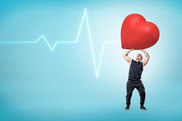 Man in sleeveless hoody holding up big heavy red heart on light-blue background with pulse line and much copy space.