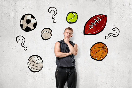 Front view of young man in sleeveless hoody standing with hand rubbing chin at wall with drawings of different sport balls and question marks among them. - Powered by Adobe