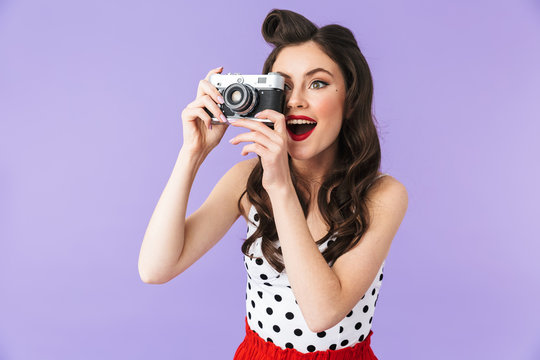 Portrait of european pin-up woman 20s in vintage polka dot dress smiling while holding retro photo camera