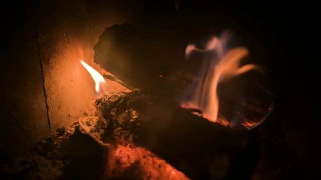 Close-up of dimly burning firewood with coals in a modern home stove behind a refractory glass at night. Low key. Movement near the fire with the camera turning. Eco-friendly energy