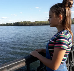 Fototapeta premium A young girl on the embankment of a large river looks at the water. Long hair gathered in a bun. Summer, striped short-sleeved clothing, jeans.