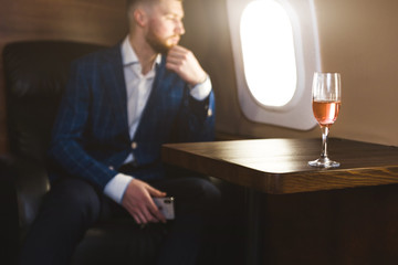 A young successful businessman in an expensive suit sits in the chair of a private jet with a glass of champagne in his hand and looks out the window