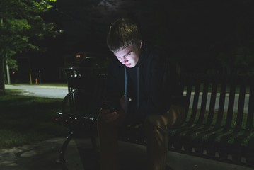 Depressed teenager on his cellphone while sitting on a park bench at night.