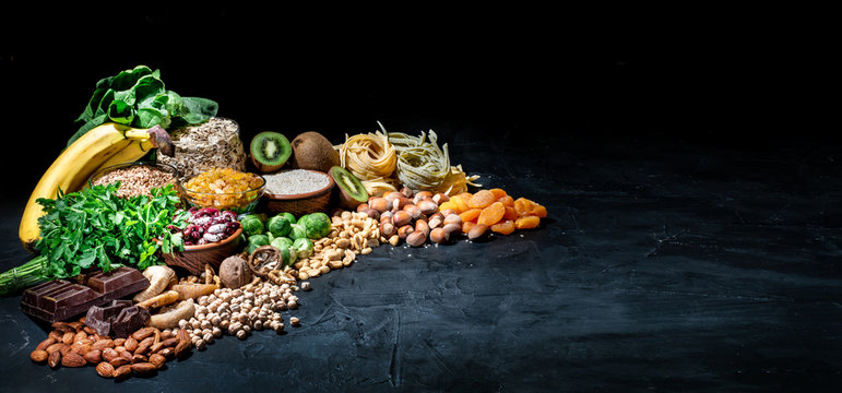 Assortment of high magnesium sources: bananas, nuts, oatmeal, buckwheat, peanuts, spinach chard, dark chocolate and sesame seeds on dark background