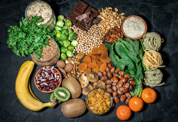 Assortment of high magnesium sources: bananas, nuts, oatmeal, buckwheat, peanuts, spinach chard, dark chocolate and sesame seeds on dark background - obrazy, fototapety, plakaty