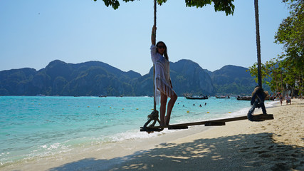 Fototapeta premium Beautiful girl in a white Cape and a black bathing suit on a swing. Swing on the beach. View of the island, beach with sand, blue water and green leaves of trees. Visible hills of the island.