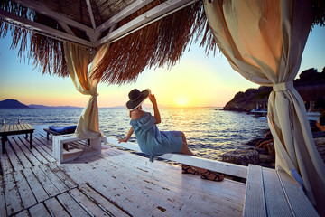 Woman in hat relaxing by the sea in a luxurious beachfront hotel resort at sunset enjoying perfect...