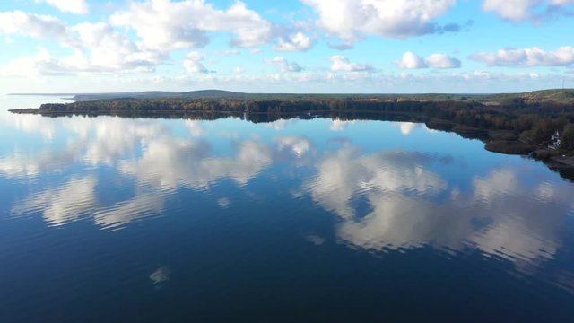 Panoramic drone shot of Pervalka, small village in Lithuania, surrounded by autumnal forest, near the coastline and the calm sea, reflecting white clouds on the surface, on a bright sunny day
