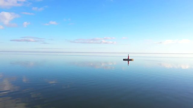 Drone shot of a small red light house with birds flying around, near Pervalka, Lithuania, near the coastline, in the calm sea with reflecting surface, on a bright sunny day