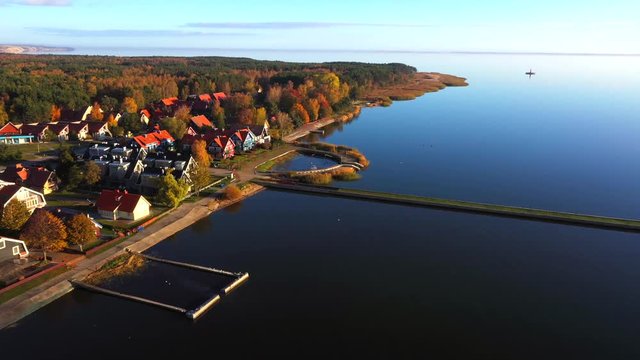 Panoramic drone shot of Pervalka, Lithuania, surrounded by autumnal forest, near the coastline and the calm sea, reflecting clouds on the surface, a lighthouse on the background, on a bright sunny day