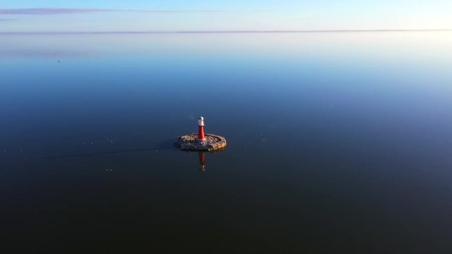 Drone shot of a small red light house with birds flying around, near Pervalka, Lithuania, near the coastline, in the calm sea with reflecting surface, on a bright sunny day
