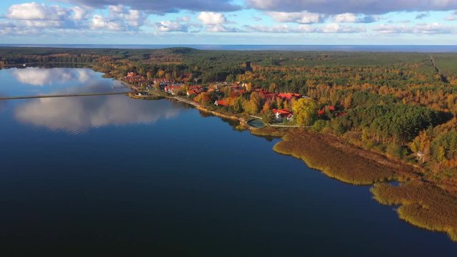 Panoramic drone shot of Pervalka, small village in Lithuania, surrounded by autumnal forest, near the coastline and the calm sea, reflecting white clouds on the surface, on a bright sunny day