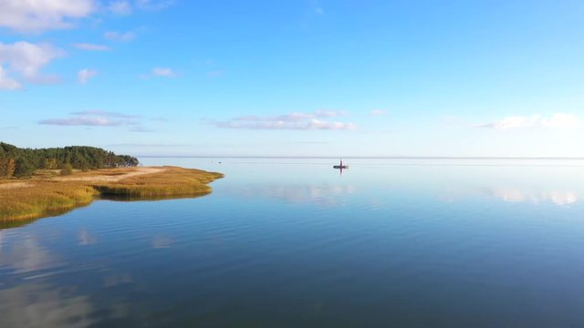 Drone shot of a small red light house with birds flying around, near Pervalka, Lithuania, near the coastline, in the calm sea with reflecting surface, on a bright sunny day