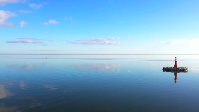 Drone shot of a small red light house with birds flying around, near Pervalka, Lithuania, near the coastline, in the calm sea with reflecting surface, on a bright sunny day